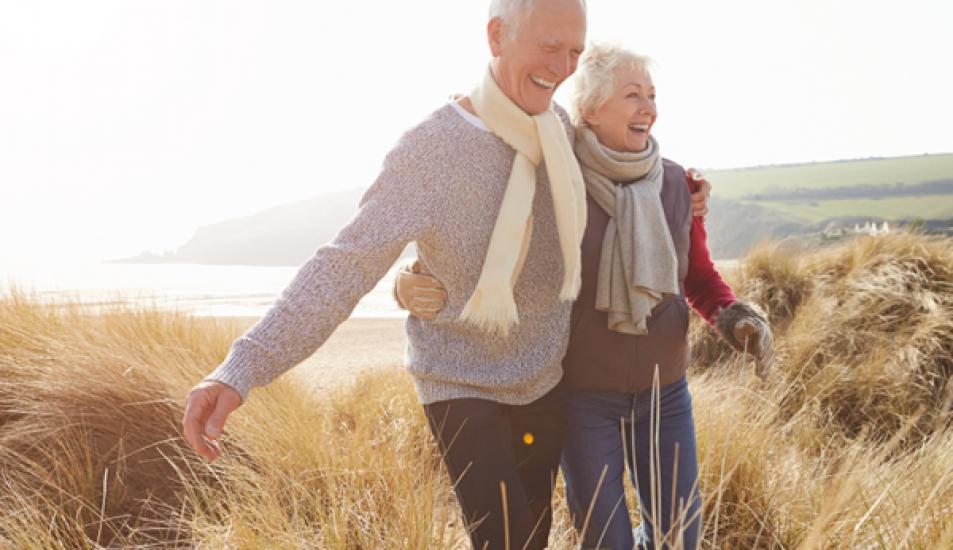 Senior Couple Walking Through Sand Dunes On Winter Beach Smiling